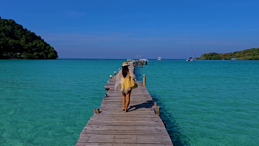 A woman strolls along a rustic wooden pier in Koh Kood Thailand, surrounded by vibrant turquoise waters and lush greenery in Thailand. The bright blue sky enhances the serene atmosphere.