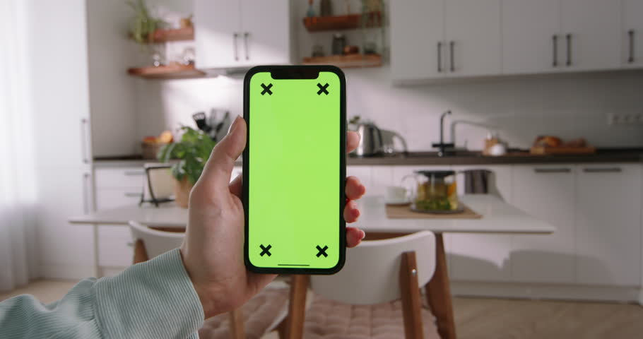 Woman holding smartphone with green screen, tapping in center, exploring various video contents on social media. Enjoying modern mobile entertainment in comfort of her living room. App advertising