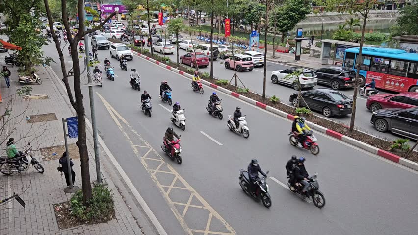 Hanoi, Vietnam Feb 7, 2025: High angle view of a busy city street lined with trees, featuring cars and motorbikes and pedestrians moving in both directions on multiple lanes.