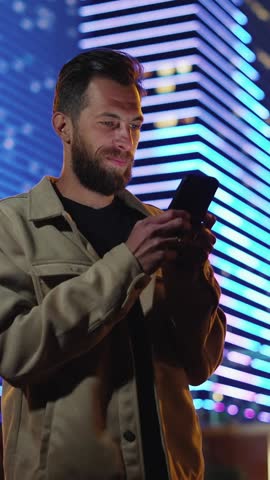 One Man with Phone in Hand at Dark Urban Street Scrolling News. Person Walks in Neon Evening and Uses Smartphone Near Futuristic Illuminated Buildings. Electronic Information for Big Life of Traveler