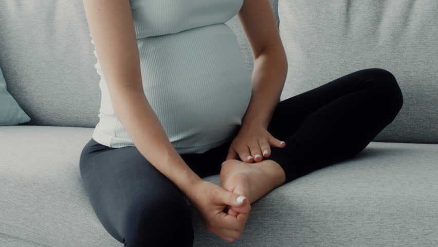 A pregnant woman sits comfortably on a cozy couch, gently cradling her baby bump. She appears relaxed and reflective, enjoying this special moment at home, massaging her feet