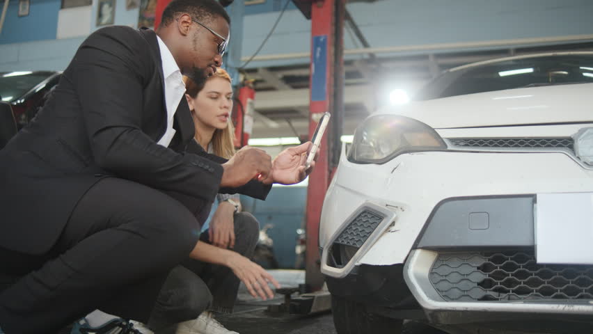 Warranty service business, Black male insurance agent inspector records crash car damage bumper, taking a photo for claim comprehensive of wreck fix list to young White female customer at auto garage.