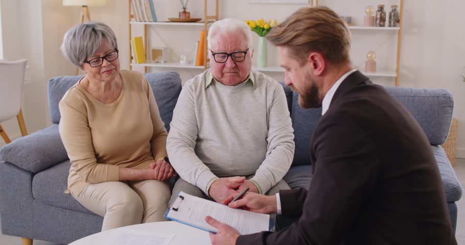 Young financial advisor in suit consulting elderly couple at home. Senior man and woman listening attentively, discussing documents, making important financial, legal or life insurance decisions.