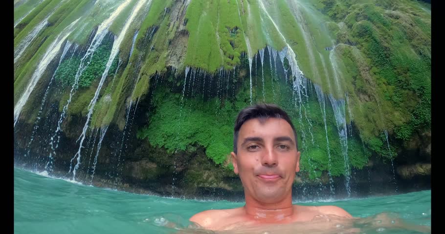 A man is swimming in a pool with a waterfall in the background. The waterfall is green and has a lot of water coming out of it. The man is smiling and enjoying the water