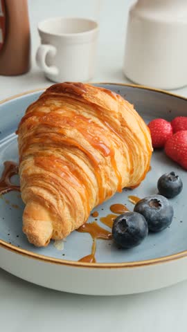 Vertical screen: pouring fruit topping on a fresh croissant in a plate. Drizzle liquid caramel over a freshly baked croissant topped with ripe berries. A nutritious breakfast or snack at work.