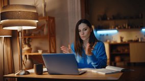 Employee consulting at virtual conference gesturing to laptop web camera in night home. Smiling woman freelancer explaining task by video call. Cheerful brunette talking at online meeting late evening - Powered by Shutterstock - Get 15% off with code: PIKWIZARD15