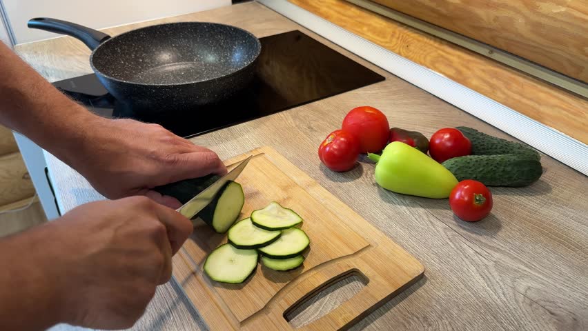 Male hands cutting green onions on chopping board at home. Fresh ingredients for food preparation - tomatoes, peppers on kitchen counter