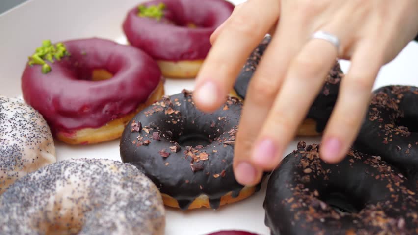 Woman's hand with manicure and wedding ring picking a chocolate glazed donut from a box full of colorful donuts with various toppings, including chocolate flakes, poppy seeds and pistachio grains