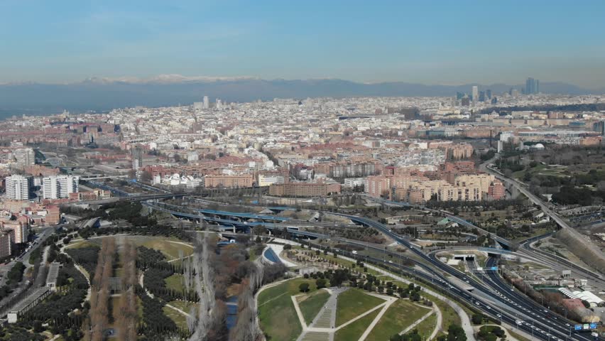 Madrid cityscape, Manzanares Park, highway interchange, traffic, buildings, distant skyline, mountains, establishing shot, sunny day, Spain. Aerial forward