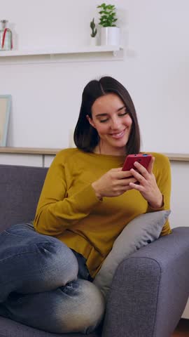 Vertical HD video of happy caucasian woman relaxing on sofa using mobile phone. Millennial female resting on couch while holding cellphone device, watching content on social media networks
