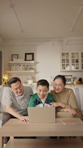 Asian boy sits on sofa with his grandparents, patiently showing them how to use a laptop in cozy home, intergenerational learning, digital literacy for seniors, family connection across generations.