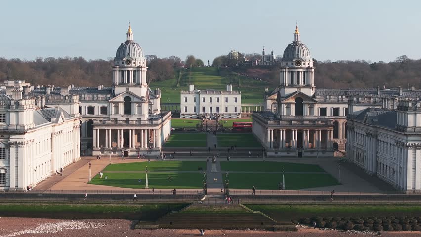 Aerial view of the Old Royal Naval College in Greenwich, London, with twin domes, Greenwich Park, Royal Observatory, and a red double decker bus in motion.