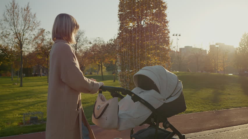 Young mother walks through the lush park with her stroller as the sun sets, creating a serene atmosphere filled with warm colors and beautiful scenery.