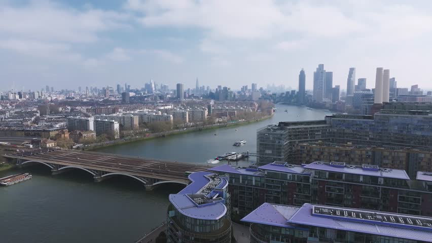 Aerial view of London with the River Thames, Battersea Power Station, a train crossing a bridge, boats on the water, and modern skyscrapers in the skyline.