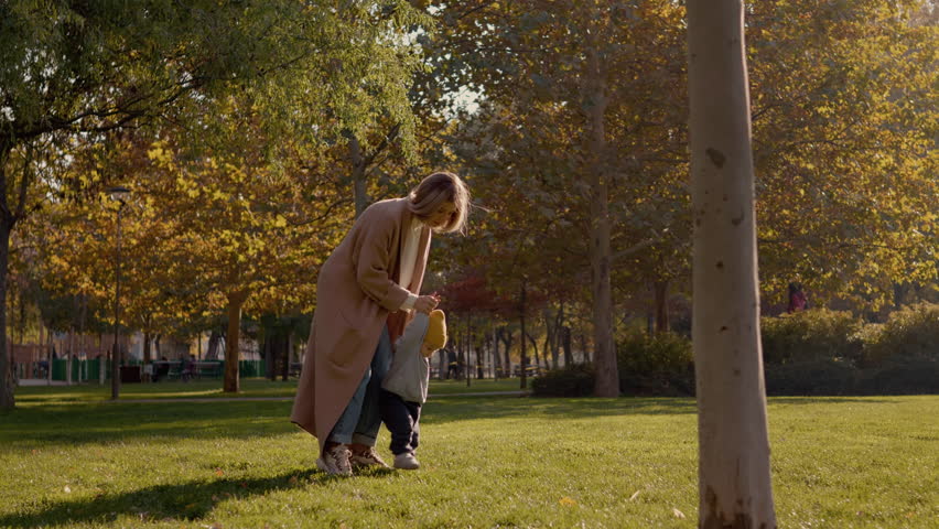 Mother encourages her young child as he takes his first steps on a lush green lawn in a park filled with autumn trees, showcasing a special bonding moment.