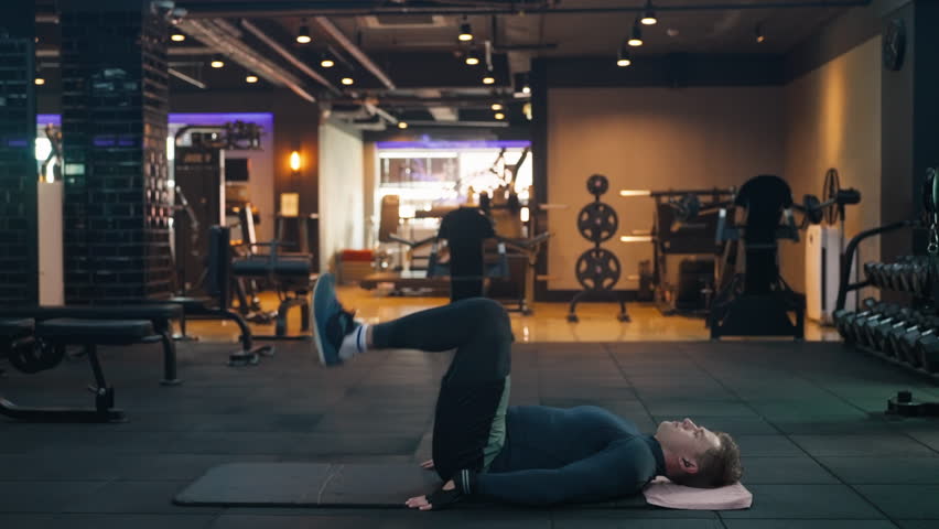 Man Lying on the Floor in Gym Doing Hip Lift Lower Abs Exercise by Pressing Feet Up Toward the Ceiling to Raise Hips Off the Ground