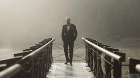 A bald man in a classic black tuxedo walks confidently across a narrow bridge surrounded by soft morning mist. He has his hands in his pockets and looks to the side with a calm, composed expression. - Powered by Shutterstock - Get 15% off with code: PIKWIZARD15