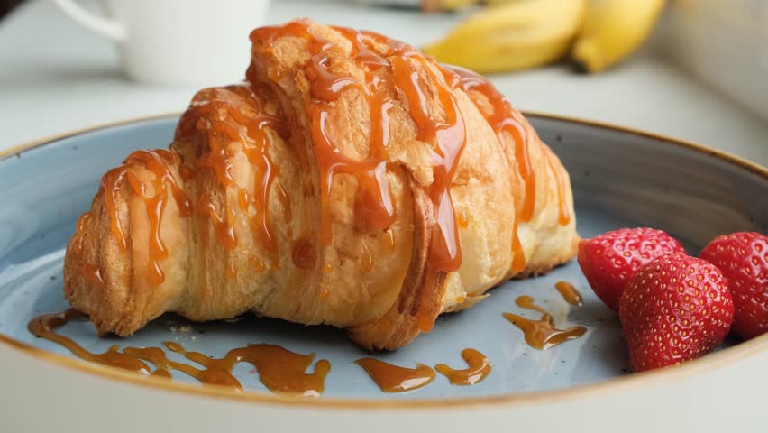 Brown shiny caramel drips from a croissant lying on a plate with strawberries. Appetizing croissant in caramel syrup. Fresh pastries are suitable for breakfast or a light snack during the working day.
