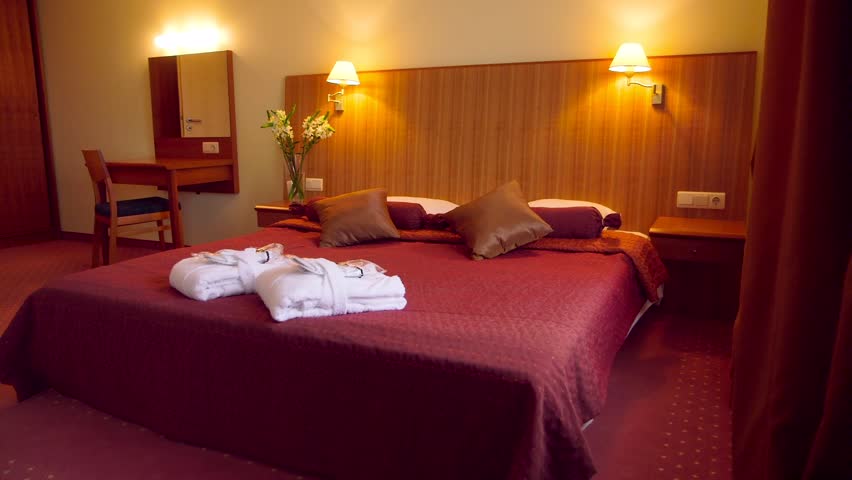 A Chic Hotel Bedroom Decorated in Red Tones. Huge Wooden Headboard. Lit Night Lights. Two White Robes and Slippers on the Bed for a SPA Visit.
