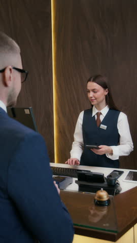 Vertical shot of hotel receptionist typing guest information on computer, then handing him passport and key card after checkin at front desk