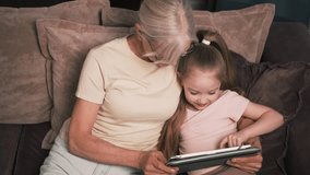 Grandmother and granddaughter sit together on the couch, reading an engaging e-book on a tablet in a cozy home setting. Reading an e-book on a tablet - Powered by Shutterstock - Get 15% off with code: PIKWIZARD15