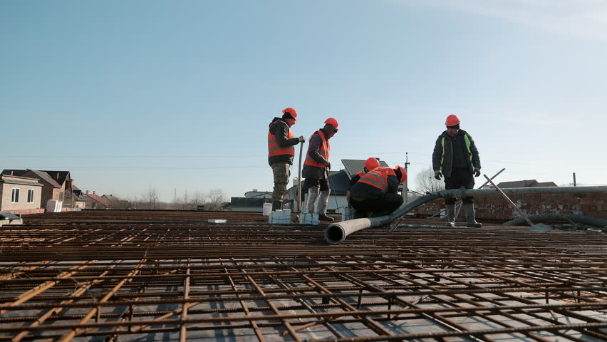 Construction workers in safety vests and helmets working with rebar and concrete hose, Outdoor building site during foundation preparation under clear blue sky