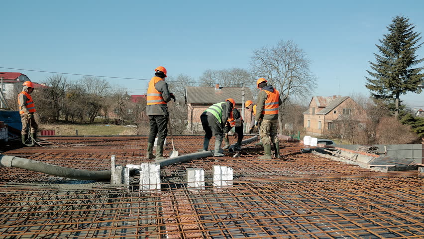Team of construction workers in orange vests pouring concrete over rebar grid outdoors, Coordinated foundation work on residential building site in rural area under blue sky