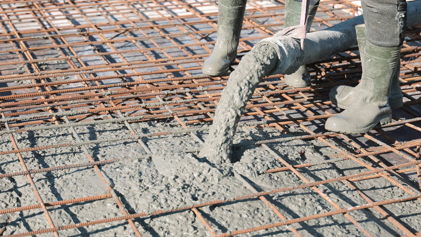 Close-up of wet concrete pouring from hose onto rebar grid with workers in rubber boots, High-detail macro of cement mix covering reinforcement for foundation construction