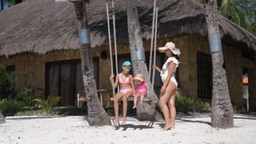 Young mother in a white swimsuit and straw hat swings her older daughter and baby daughter on a tropical beach, surrounded by palm trees and white sand. Slow motion - Powered by Shutterstock - Get 15% off with code: PIKWIZARD15