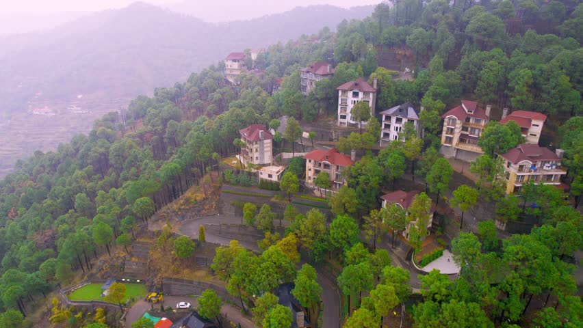 aerial drone shot panning up reveal going from houses on the side of winding road surrounded by lush green trees revealing fog covered Himalaya mountains in distance in Kasauli