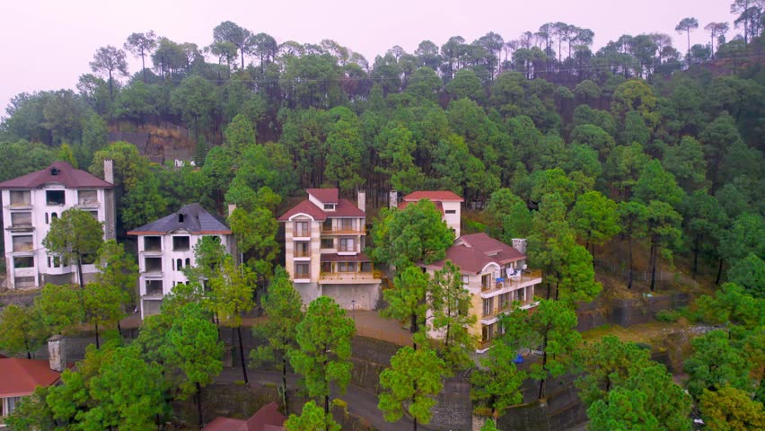 aerial drone panning shot flying backward reveal of houses surrounded by trees on hillside in Kasauli, Manali, Shimla India with fog covered mountains in the distance on a cloudy monsoon day