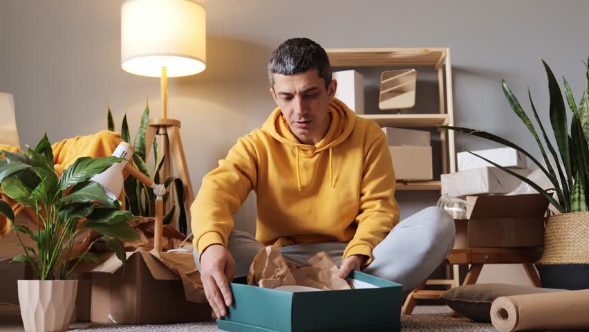 A person sits comfortably on the floor of their new living room, unpacking a box filled with items. Surrounding them are various packed boxes and houseplants, creating a cozy atmosphere