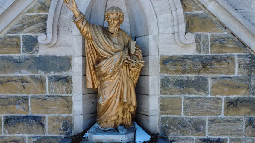 Aerial shot moving away from a gilded statue of Saint Peter embedded between a pair of bell towers on a brick facade with large stained glass windows of the Saint-Pierre-du-Lac Church, Val-Brillant. - Powered by Shutterstock - Get 15% off with code: PIKWIZARD15