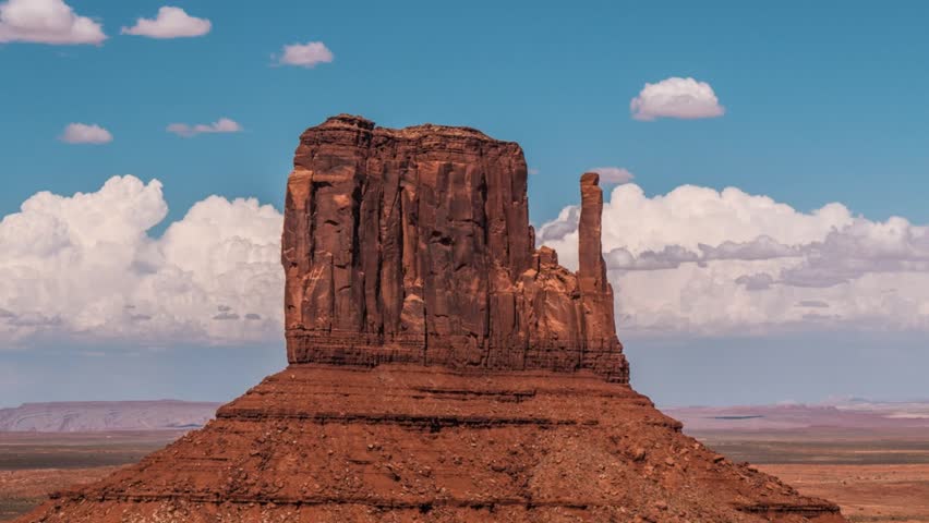Timelapse: Clouds drift over iconic Mitten butte in Monument Valley, Arizona Utah. Majestic red rock desert landscape under vibrant blue sky. Famous American West landmark.