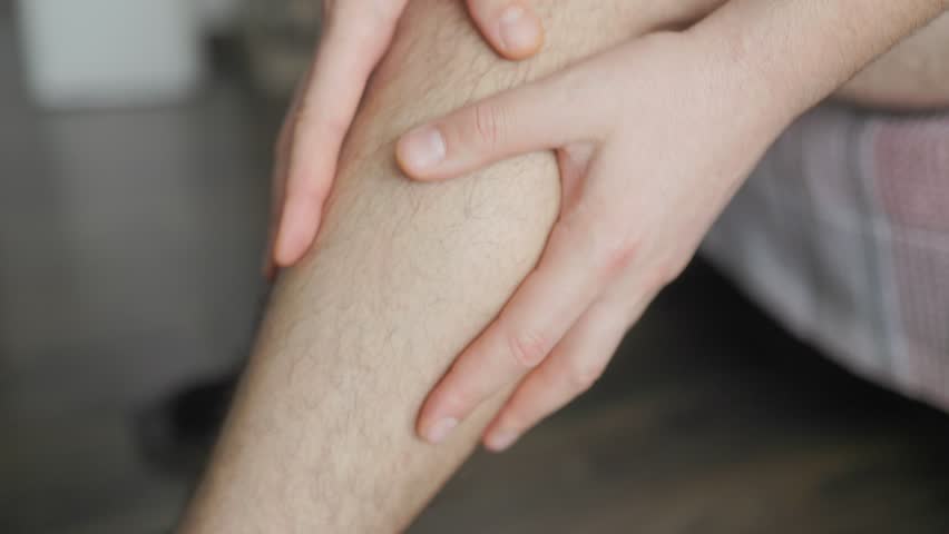 Close-up of a man rubbing his lower leg with both hands, possibly due to pain, fatigue, or discomfort. The image shows detail of skin texture and body hair.