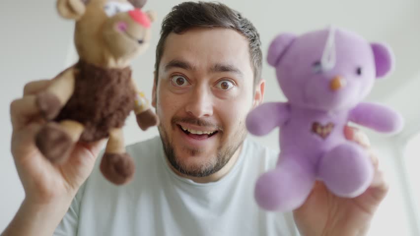 Bottom view of a cheerful man holding a purple teddy bear and a reindeer plush toy, looking at the camera and pretending to play with a baby in a bright room.