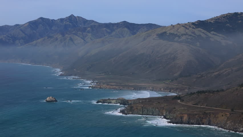 This aerial view captures Californias coastline, showcasing Big Sur and iconic Highway 1. Majestic mountains meet crashing waves at sunset, highlighting the areas stunning natural beauty