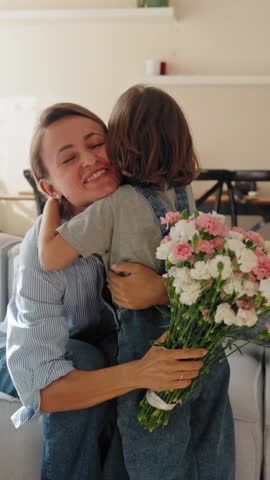Young boy gives his mother a bouquet of flowers at home in a warm and cozy setting. She smiles with joy and gratitude. Concept of Mother's Day celebration and family love. Mid-range shot natural light