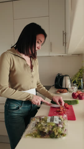 Vertical video, Young woman makes a healthy salad in a modern kitchen. She slices fresh vegetables. The focus is on wellbeing and healthy eating.