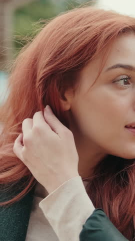Vertical video, Beautiful redhead woman smiles at the camera. She has long, red hair and is wearing a dark green jacket with a light-colored shirt.