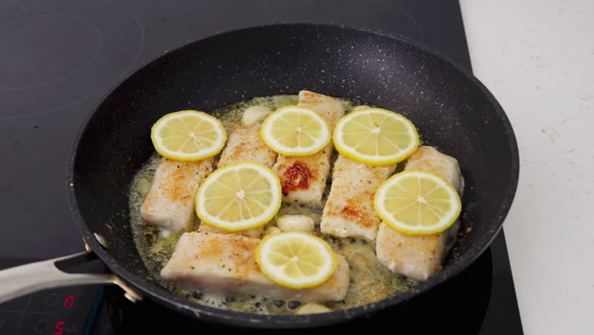  Hake fillets cook in a pan with garlic, lemon slices, and a buttery sauce.