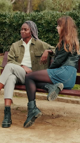 Vertical video, Two women, either a lesbian couple or best friends, sit and talk on a park bench during autumn or winter in the city.