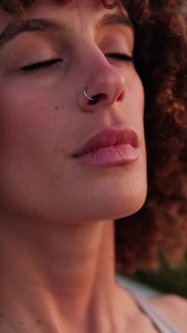 Vertical video, Young woman with curly hair breathing deeply with closed eyes. She's practicing mindfulness outdoors in sportswear, enjoying a peaceful moment.