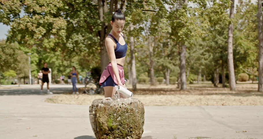 Young woman filling up her water bottle after an intense workout in the park. She is taking a break and refreshing herself.