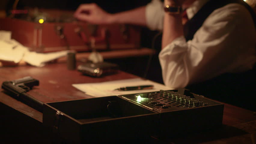 A WWII spy in a dimly lit room secretly transmits and receives messages in Morse code using a radio transmitter built into a suitcase and documents the codes.