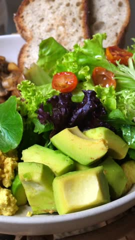 Vertical screen healthy vegan breakfast bowl featuring scrambled tofu, avocado, fresh greens, cherry tomatoes, mushrooms, and whole wheat toast gets a piece of avocado lifted by a fork
