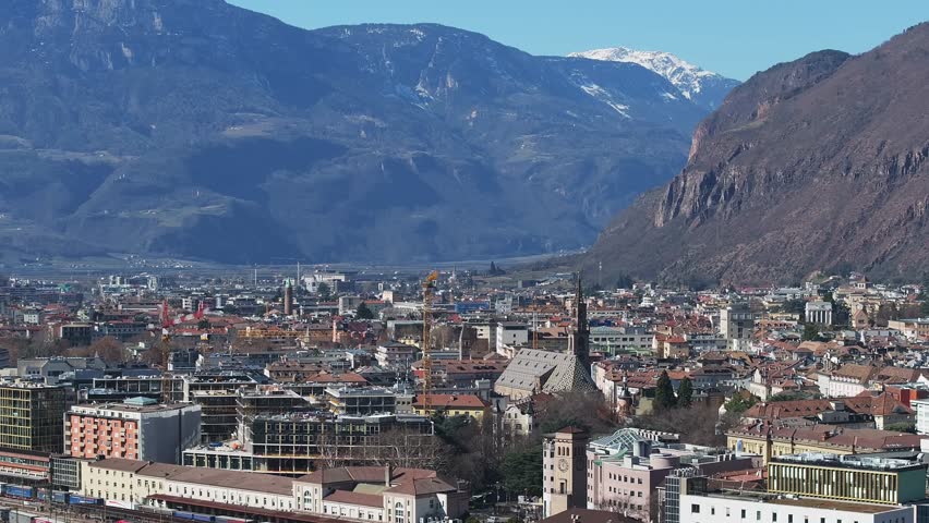 Aerial view of Bolzano, Italy, showing the Dolomites, urban architecture, a winding river, and construction areas with slow panning motion.