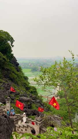Ninh Binh Vietnam - April 30 2024: vertical video of people and tourist walking up and down the Ngoa Long Mountain framed by lush greenery and Vietnamese flags outdoor at daytime in Vietnam.