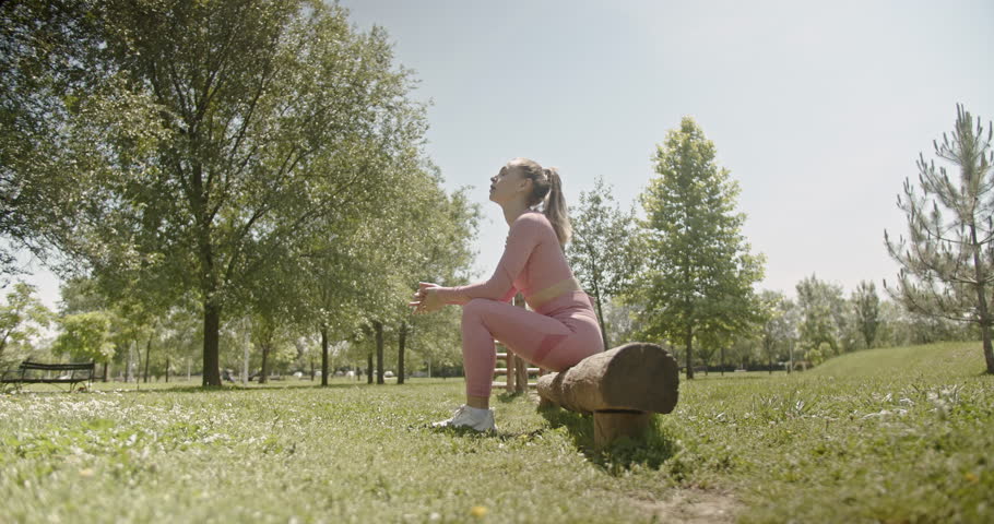 Young woman rests on a wooden beam in a park during her calisthenics workout. She