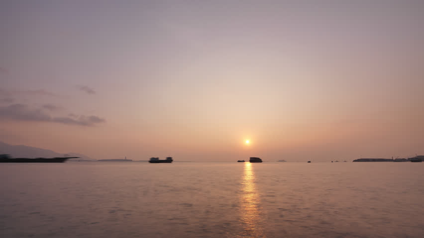 Nice sunset over waters of Urmston Road waterway, lively with container and cargo ship traffic. Wide angle timelapse shot, silhouetted vessels quickly move along harbor, sun move down and hide